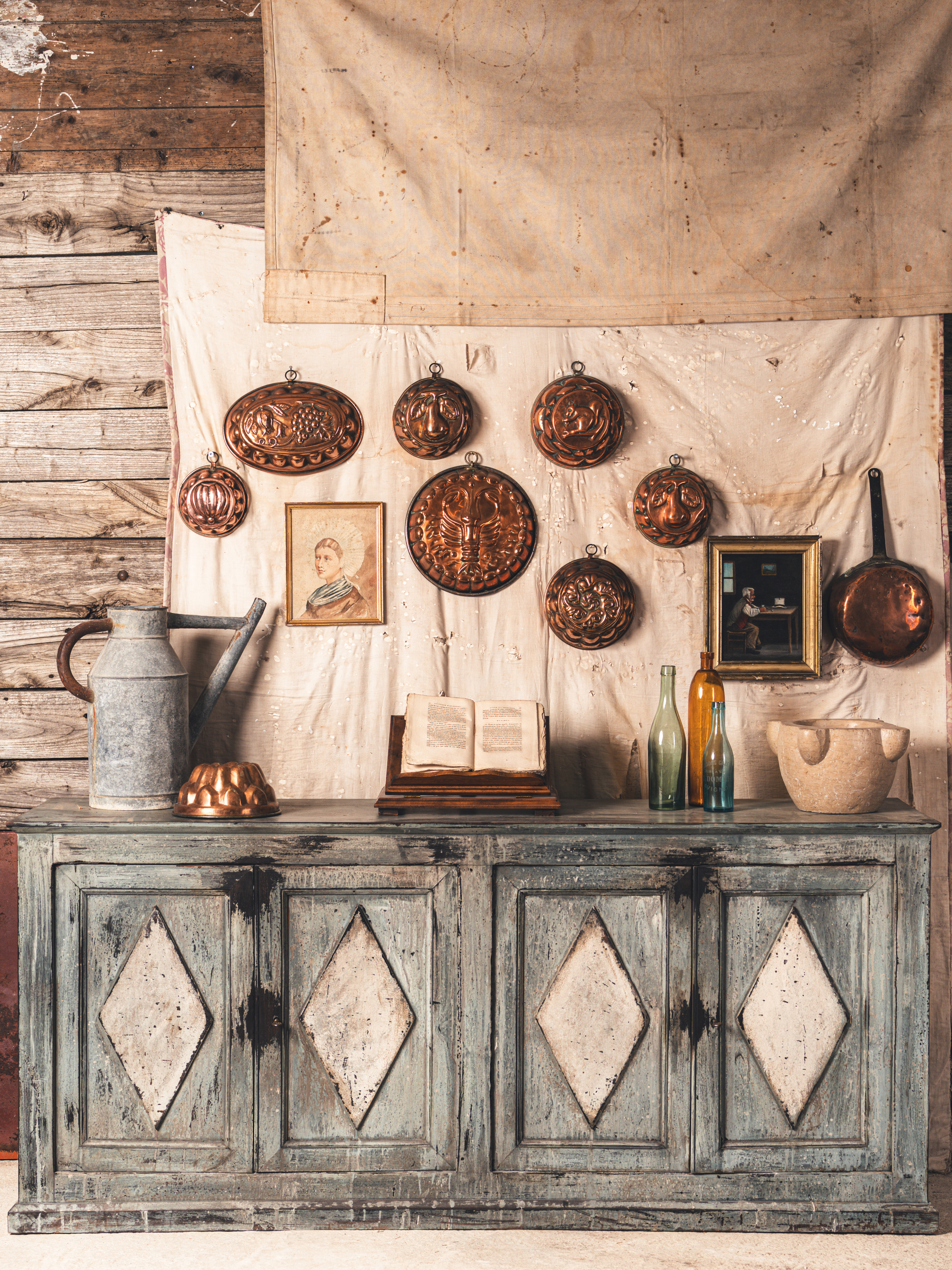 Image of an antique console cabinet painted in a light blue, weathered paint shade. The walls are an antique wood with cloths hanging on the wall. There are other antiques on the wall like copper pots, artwork, vases, etc.