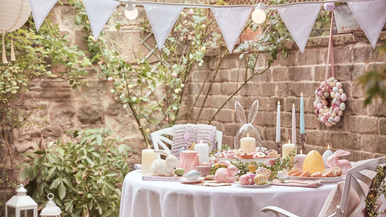 Outdoor Easter decor ideas are so cute. Here is an outdoor picnic area with blue bunting, lights, a circular dining table with a white tablecloth a spread of pastel candles, eggs, and breads and cakes, and four white iron chairs with pink pillows