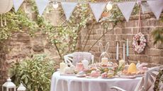 Outdoor Easter decor ideas are so cute. Here is an outdoor picnic area with blue bunting, lights, a circular dining table with a white tablecloth a spread of pastel candles, eggs, and breads and cakes, and four white iron chairs with pink pillows