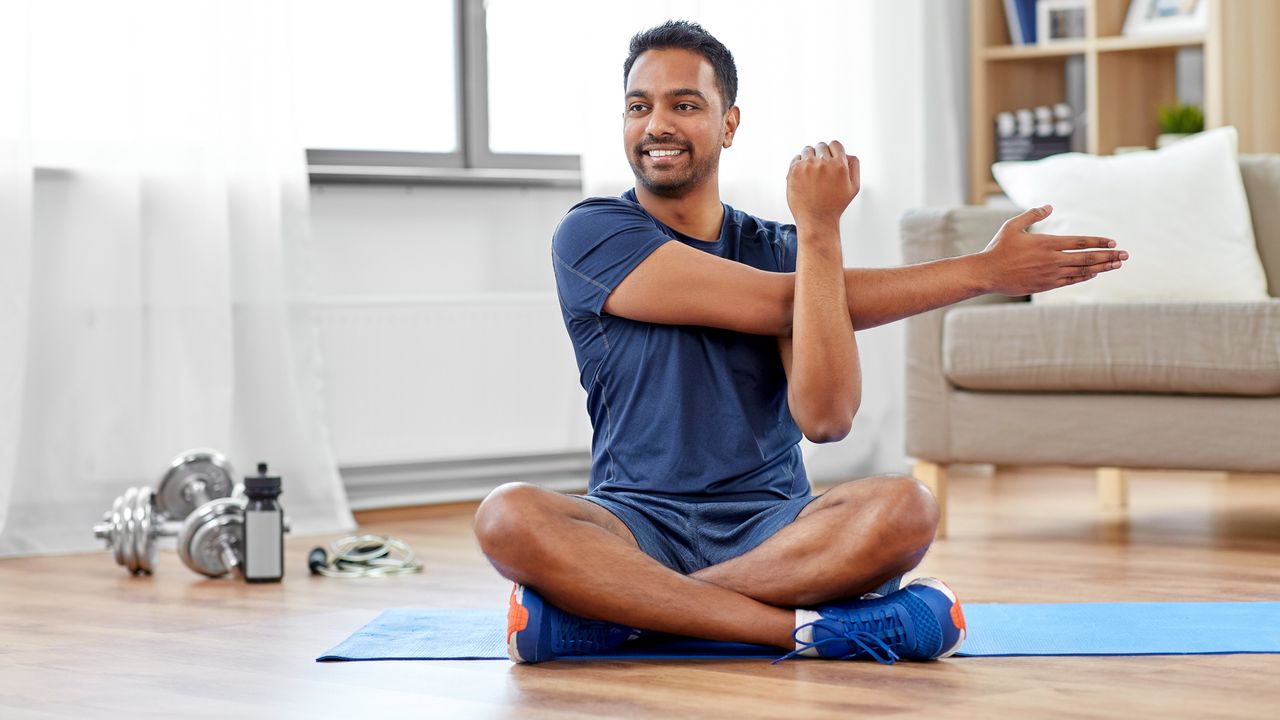 Man sitting on a yoga mat in his living room performing a stretching exercise and smiling