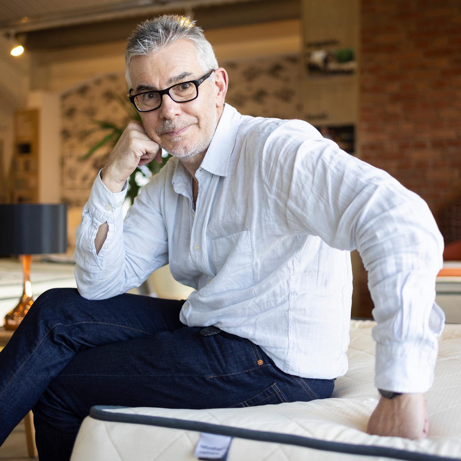 A headshot of a man with short salt and pepper hair wearing black glasses in a white shirt and blue jeans on a mattress