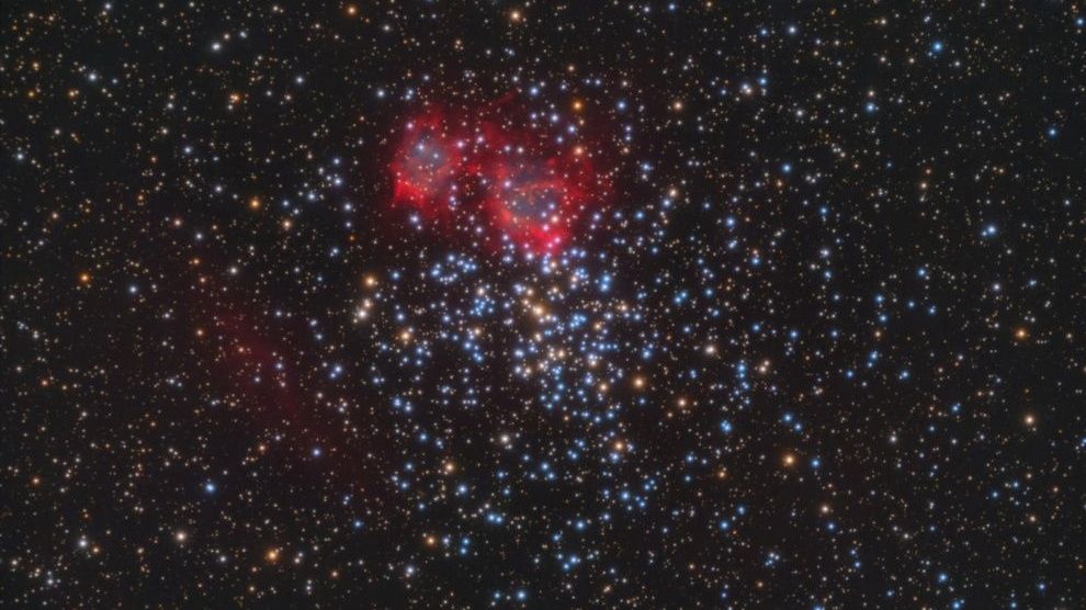 A dark sky has thick dots of stars all over it. Toward the center-top of the image, there is a pinkish-red structure that looks like dense gas. It stands out quite a bit.