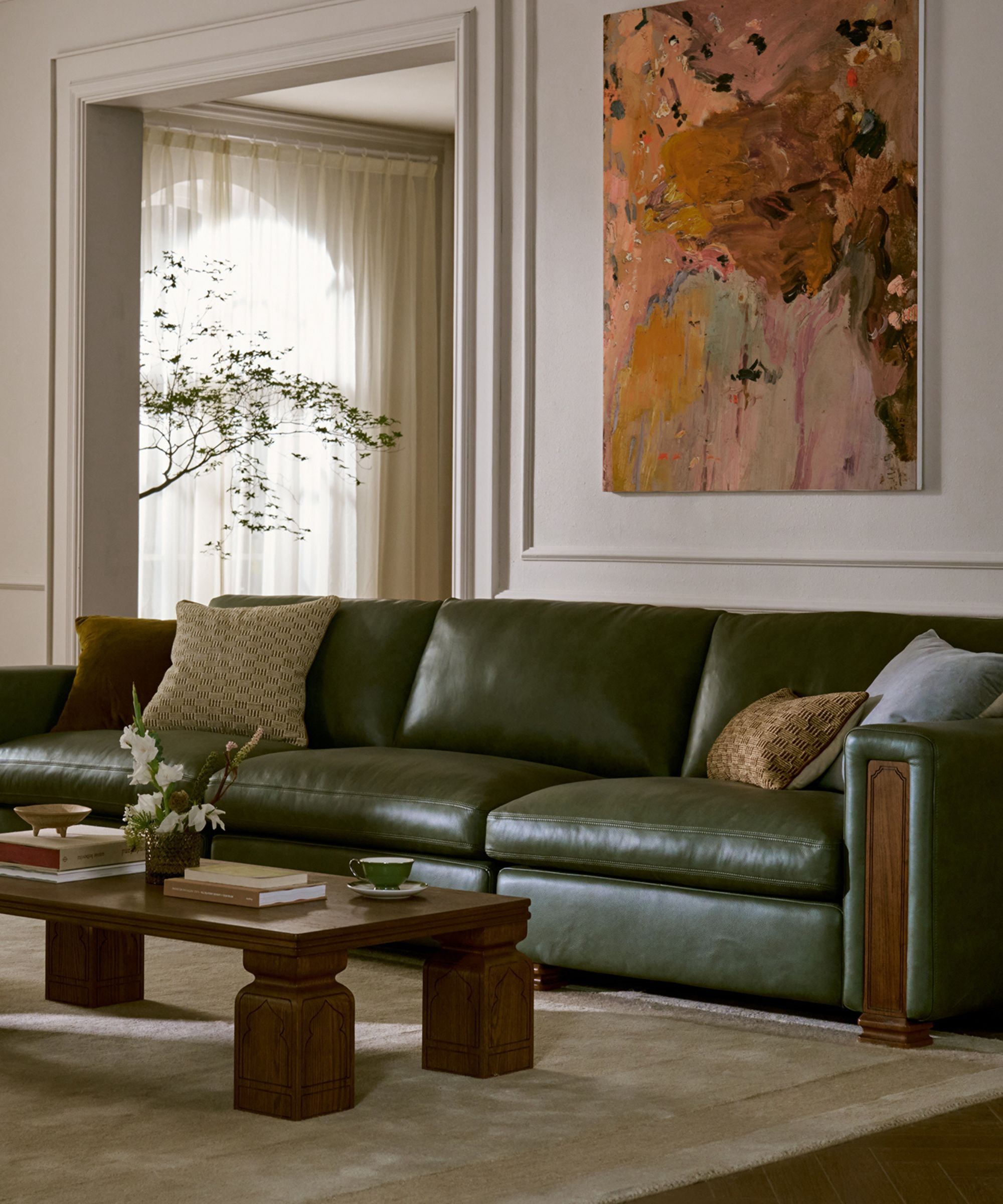 a living room with a glossy green sofa with timber detailing, colorful artwork above, beige rug, timber coffee table styled with books, and a vase of flowers