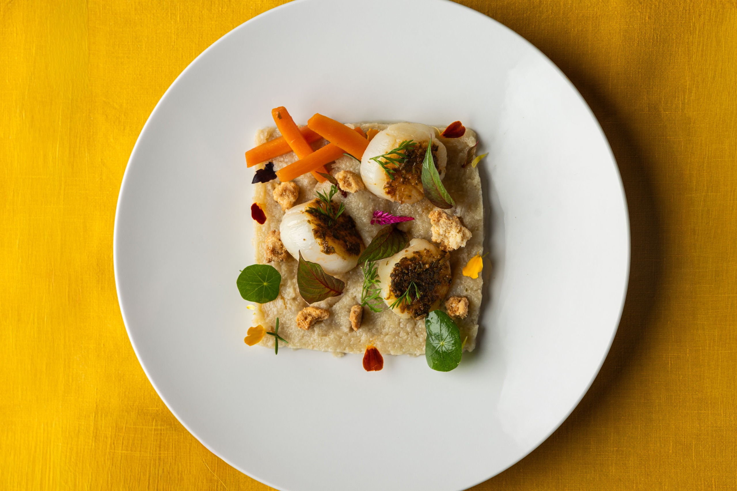 A colorful, fine dining plate of vegetables, served on a white ceramic dish atop an ochre tablecloth.