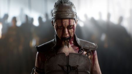 Screenshot from the Senua's Saga: Hellblade 2 cinematic trailer showing a close up of Senua staring at the camera with face markings and a blurry crowd of people behind her.