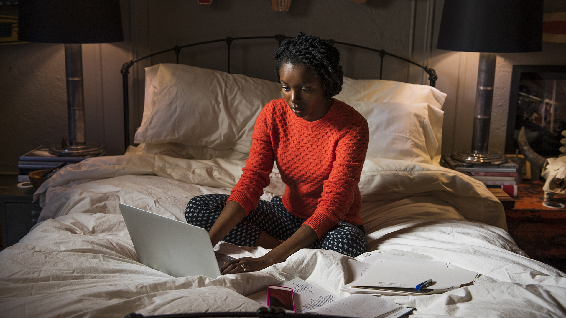 A woman sits on a bed in a dark room, typing on a laptop
