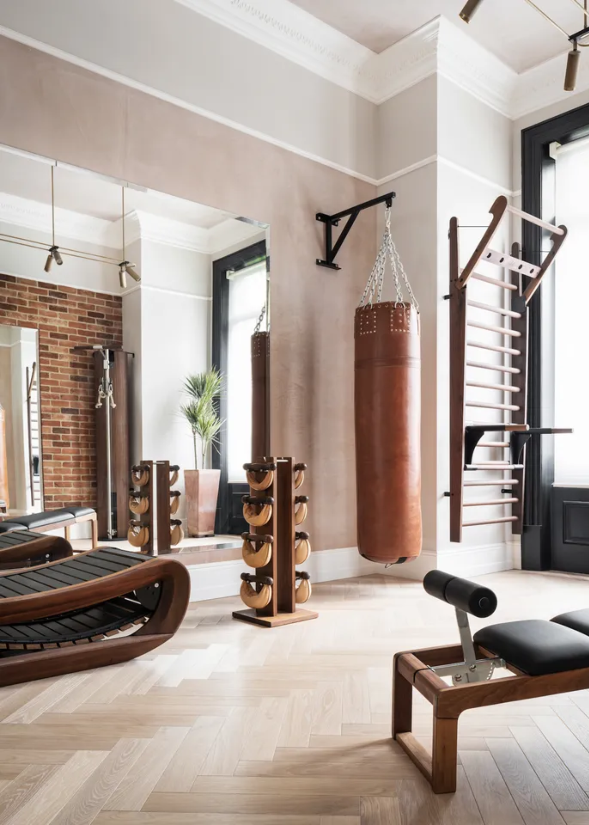 A home gym with herringbone wood floors, wood and black gym equipment including a stand of kettlebells, a bench, a punching bag, and a recliner