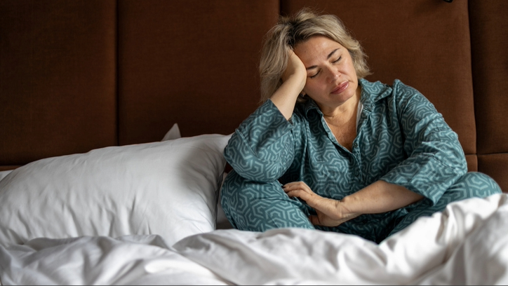 A woman wearing blue pyjamas sitting with her legs crossed and head leaning on hand in bed with white bedding against brown headboard