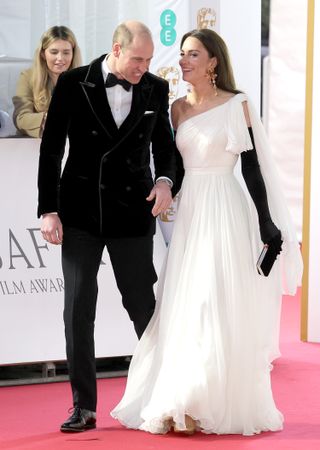 Prince William wearing a black tuxedo and holding hands with Kate Middleton in a white one-shouldered gown on the BAFTAs red carpet