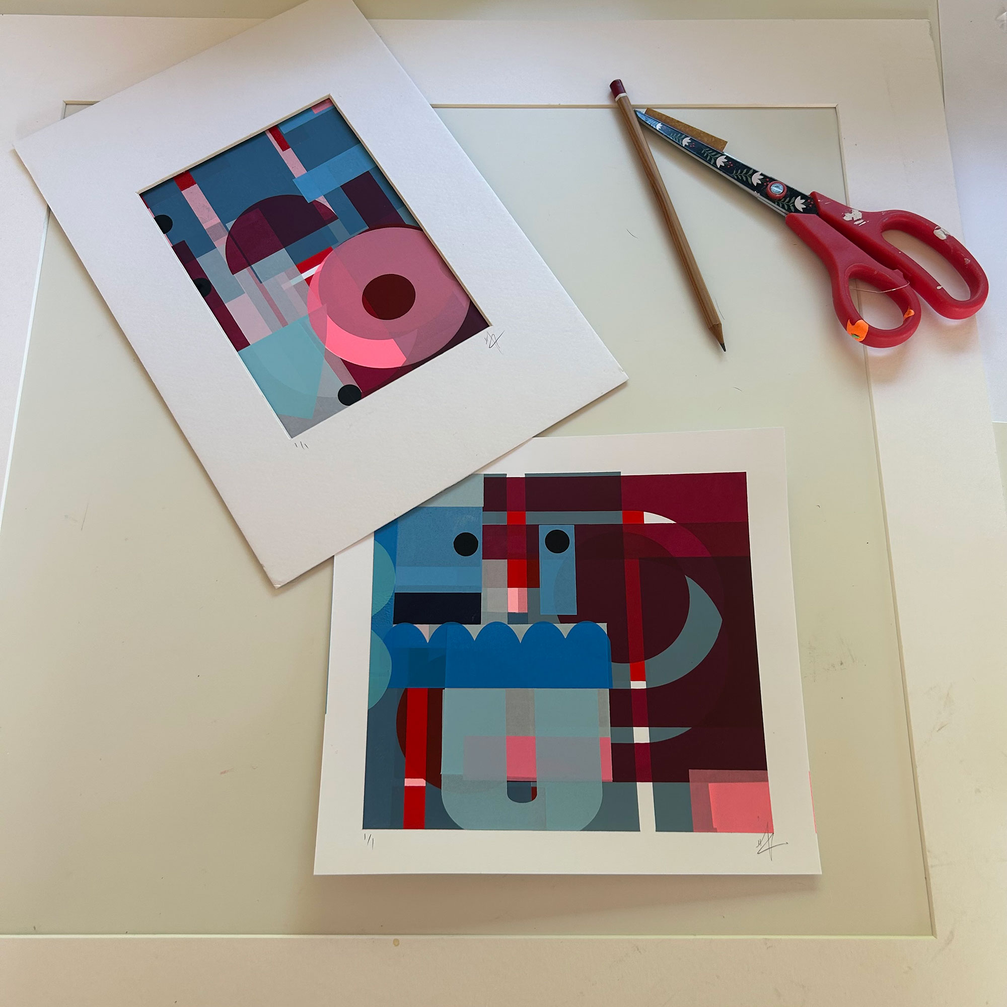 Two screenprints of blue and red shapes resting on table beside pencil and pair of scissors