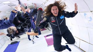 a floating participant on a zero-g airplane flight stretches out their hands and smiles in front of a crowd of people floating in all directions