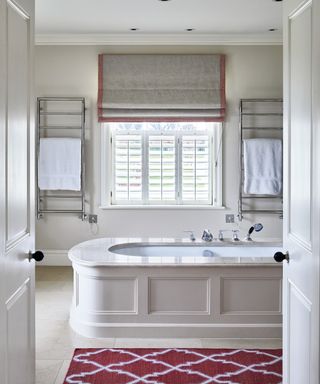 A large bath in the center of an off-white bathroom with a red and white floor rug