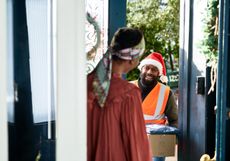 A delivery man wearing a santa hat delivers gifts to a woman at home.