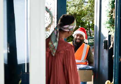 A delivery man wearing a santa hat delivers gifts to a woman at home.