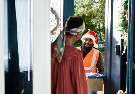 A delivery man wearing a santa hat delivers gifts to a woman at home.