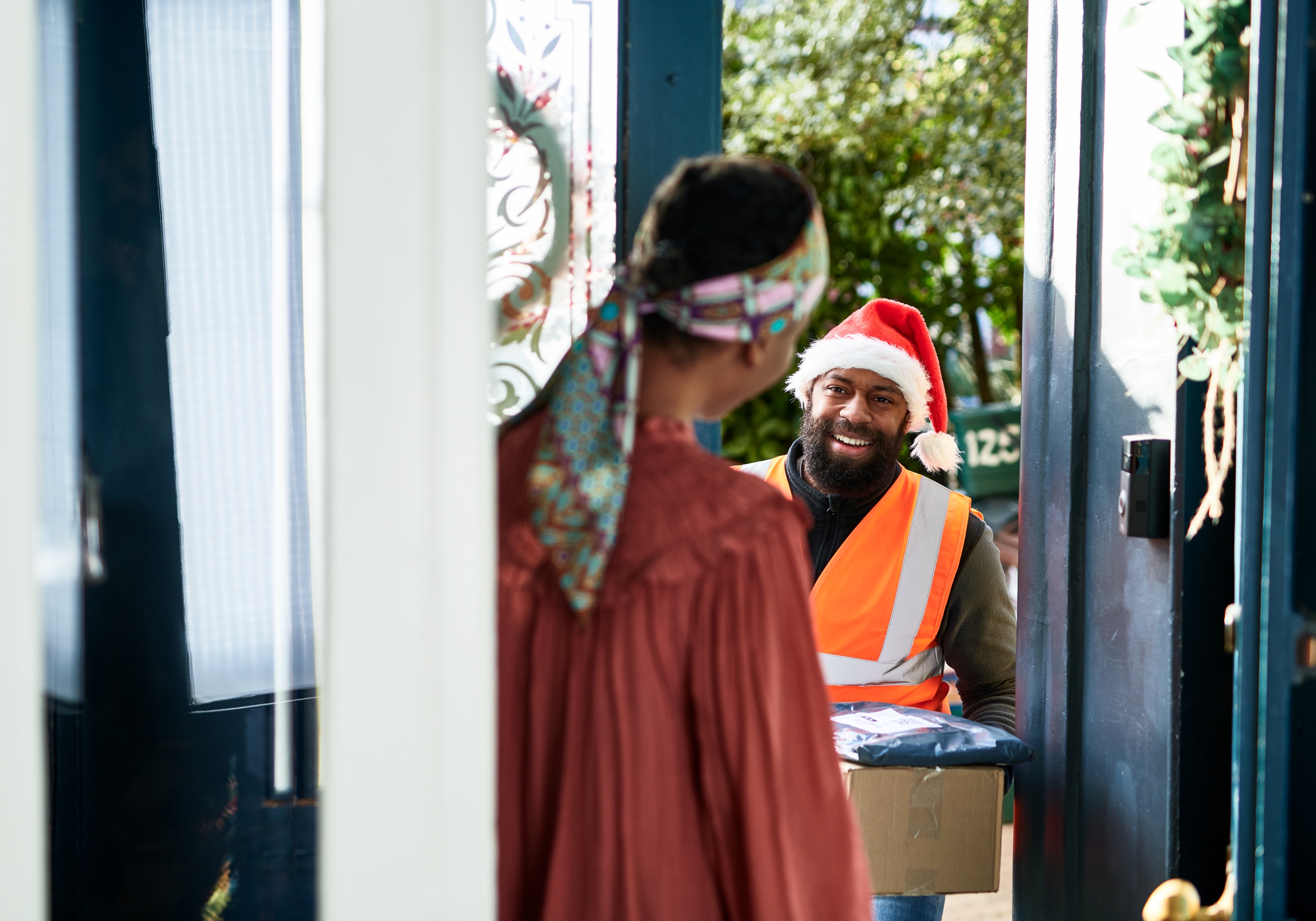 A delivery man wearing a santa hat delivers gifts to a woman at home.