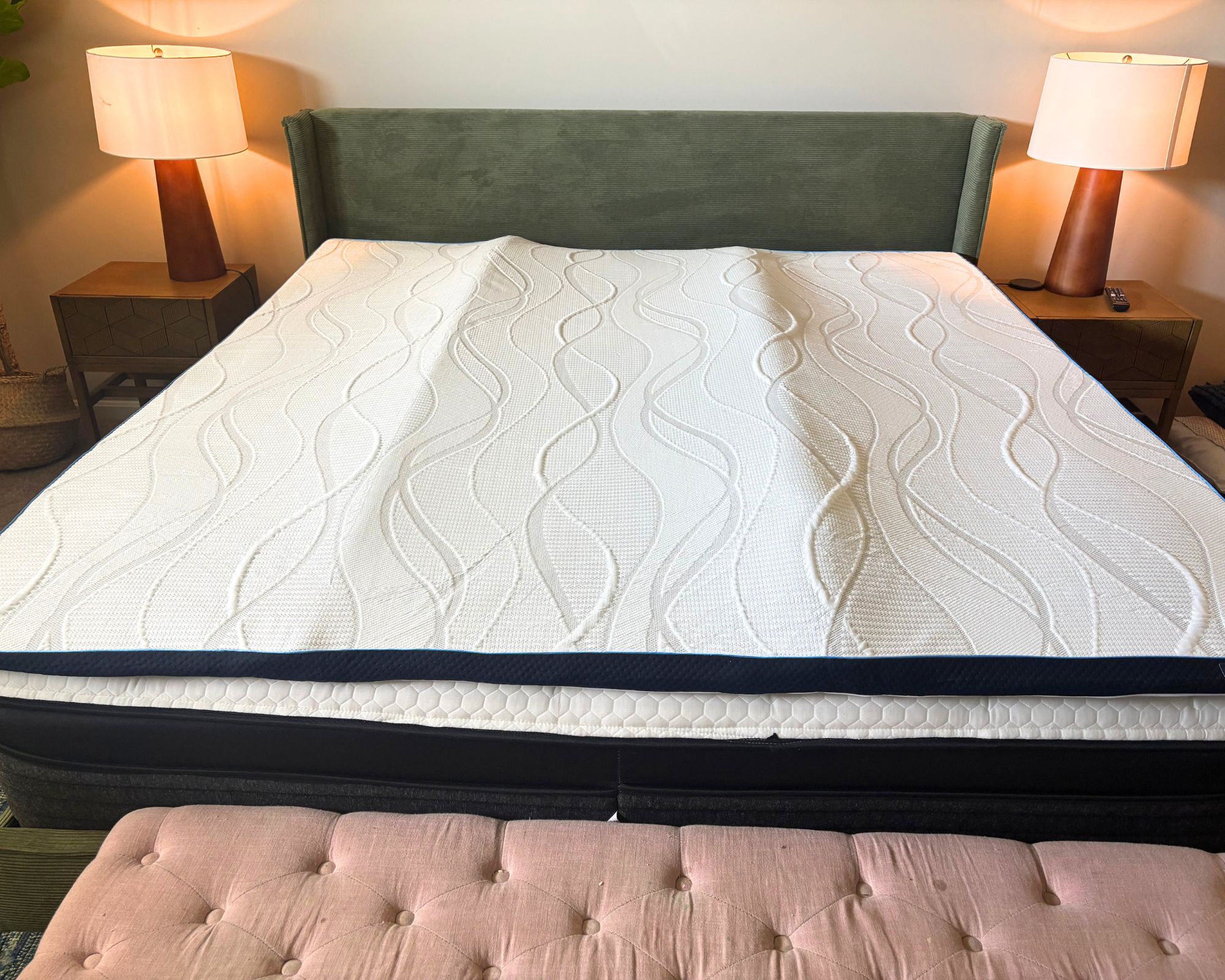 Nectar firmer memory foam mattress topper on a bed with green velvet headboard, pink ottoman, and two matching nightstands with matching brown lamps with white lampshades.