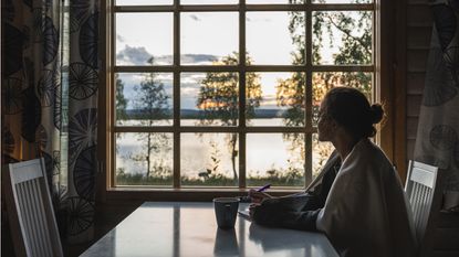 A woman sits at a table with a pen and notepad and gazes out the window at a lake view.