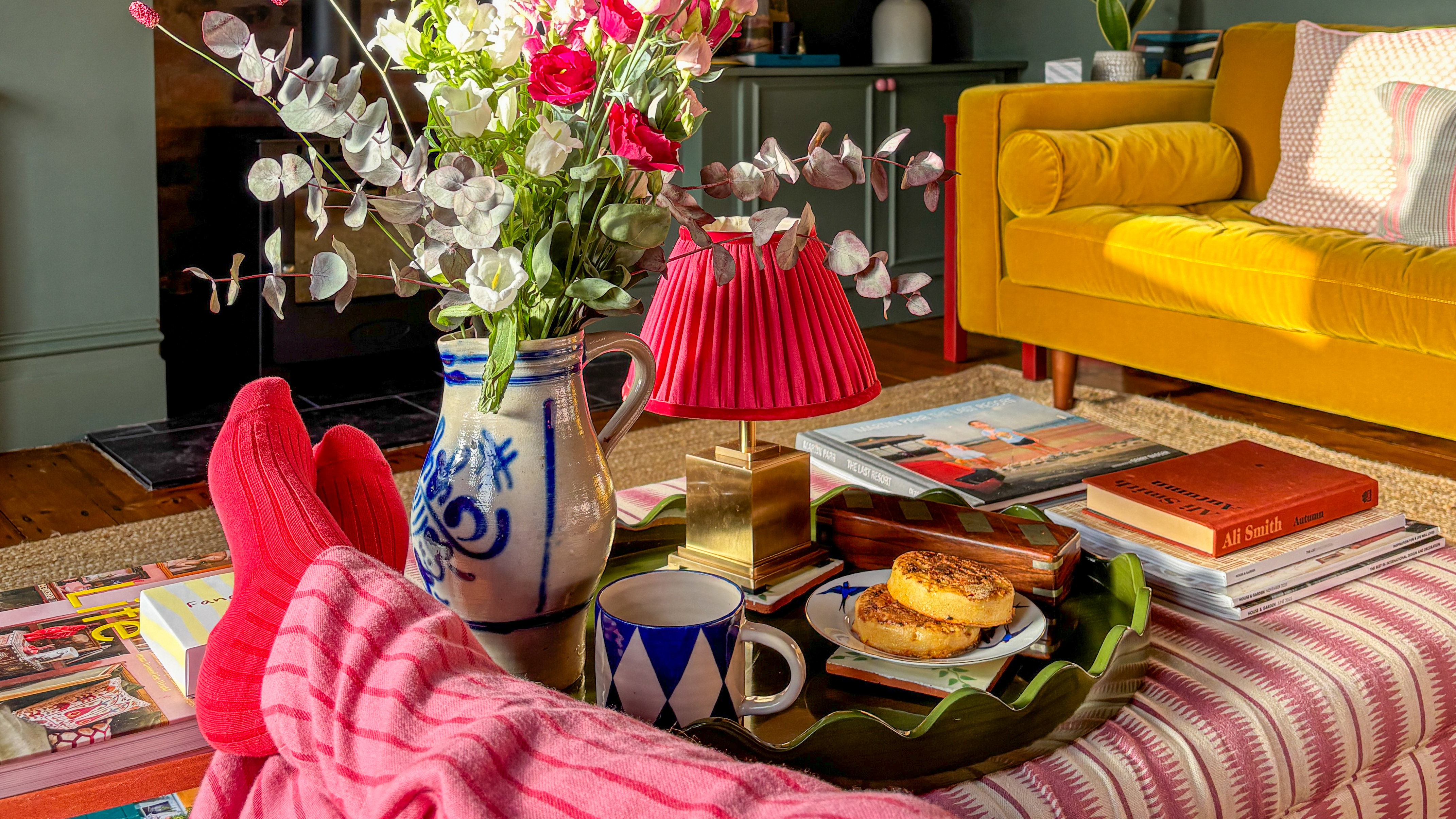 Green ceramic tray laden with a brass lamp, white and blue mug and a plate with crumpets placed on a red and white striped footstool next to a jug of flowers