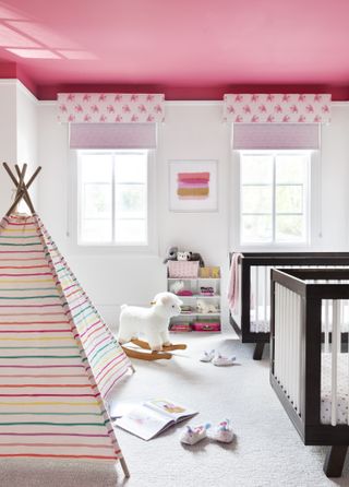 baby girl nursery with white walls, pink painted ceiling, pink print blinds, teepee, two black cots