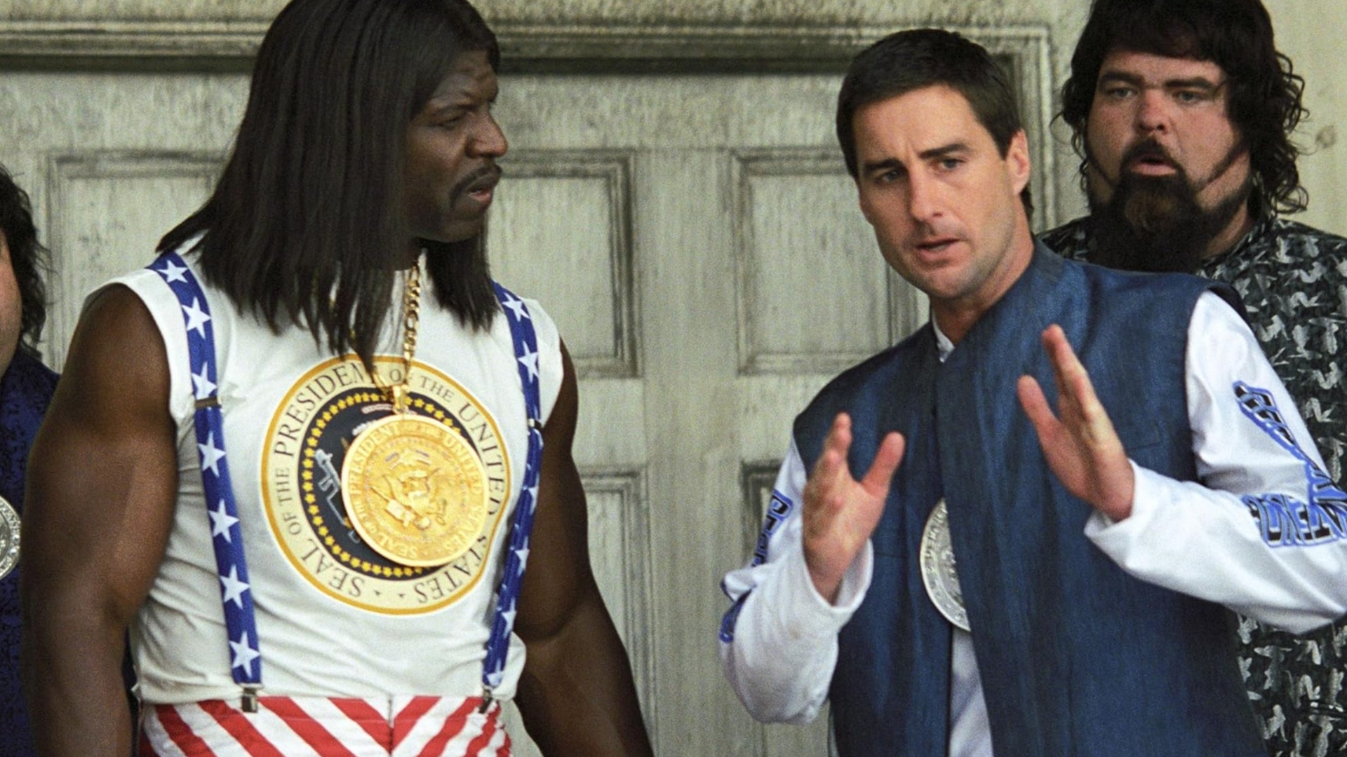 Screenshot from the movie Idiocracy. A man in a blue and white jacket is explaining something to another man who is wearing a vest with the President of the United States seal on.