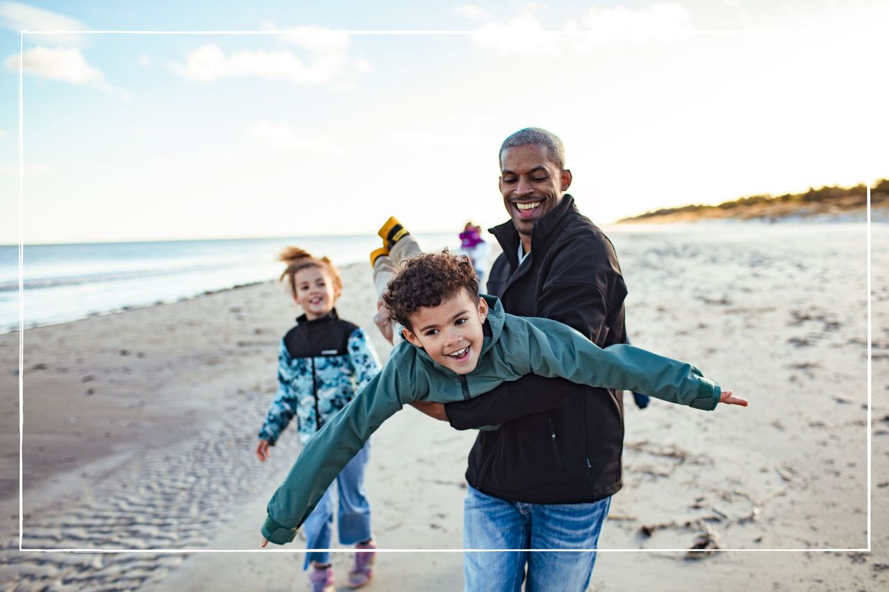 Father holding his child pretending to be an aeroplane while other kids watch on the beach