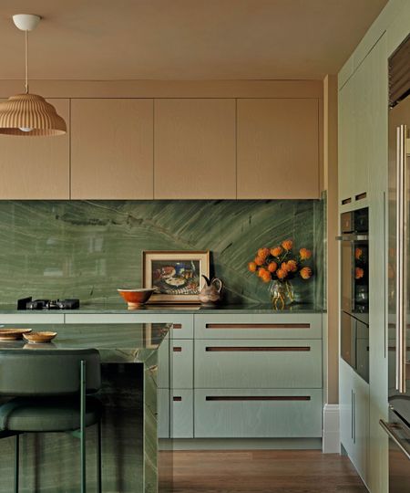 A contemporary kitchen showcasing sage green cabinetry with integrated wooden handles and a striking green marble backsplash and island. A fluted terracotta pendant light hangs above the workspace.