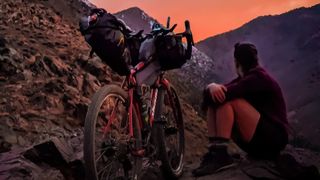 Jenny Tough is sat next to her bike looking towards an atmospheric sunset