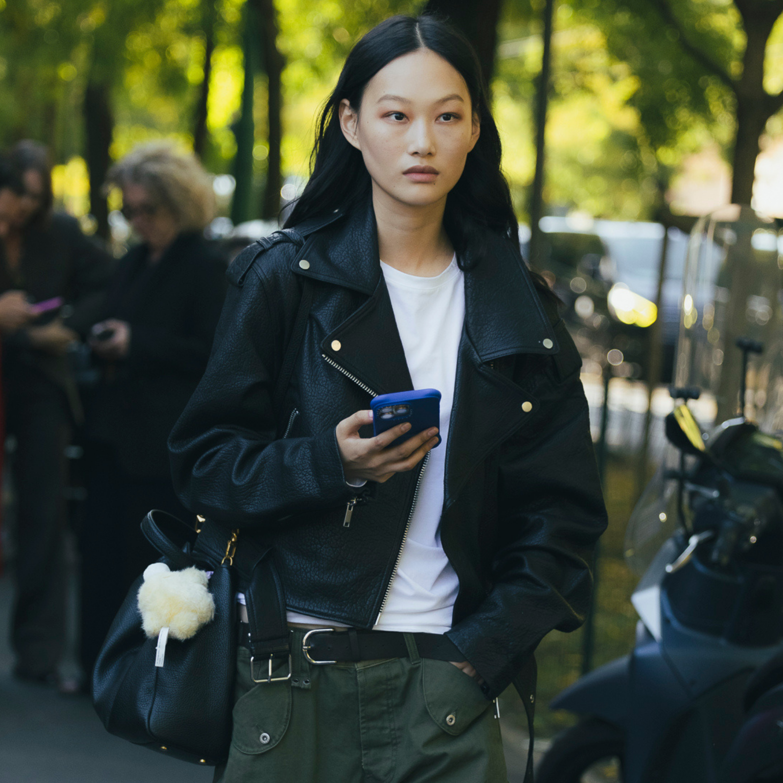 A model wears a black leather jacket with a shite shirt carrying a blue cell phone. 