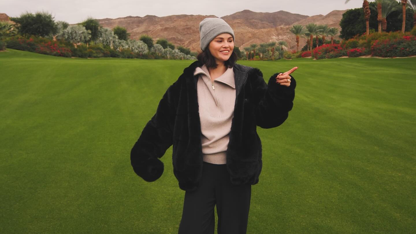 Selena Gomez wearing a beanie and black jacket, standing in a field