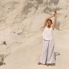 Woman stood in desert stretching and wearing white top, striped linen trousers, and brown sandals.