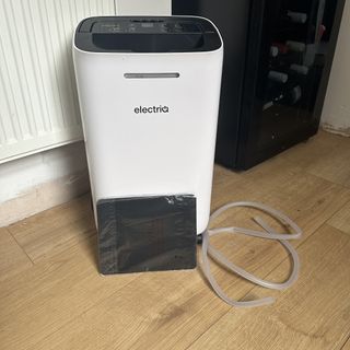 The ElectriQ 12L dehumidifier on a wooden floor with a clear drainage hose and carbon filter leant against it
