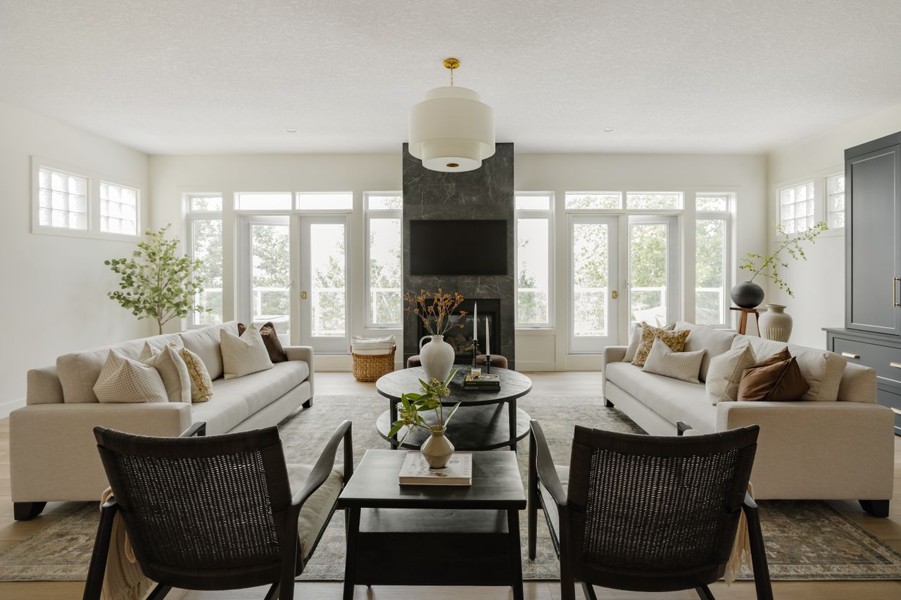 A neutral living room with white walls, two large cream couches facing each other, and two blak accent chairs