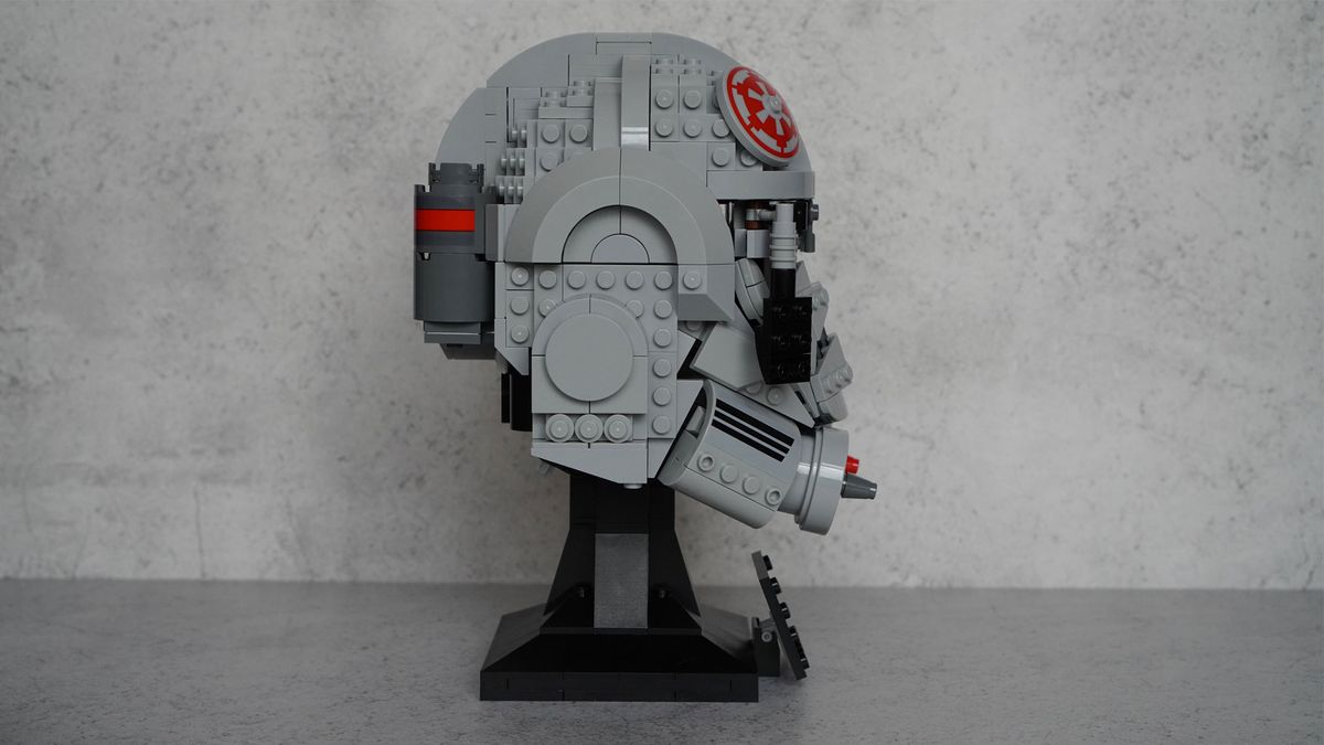 A gray helmet with black eyes and frontal features also has splashes of red on the top and the bottom is on a black display stand with a name plaque in front of a gray background