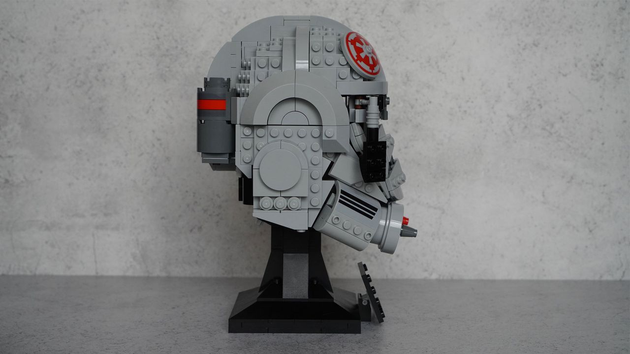 A gray helmet with black eyes and frontal features also has splashes of red on the top and the bottom is on a black display stand with a name plaque in front of a gray background