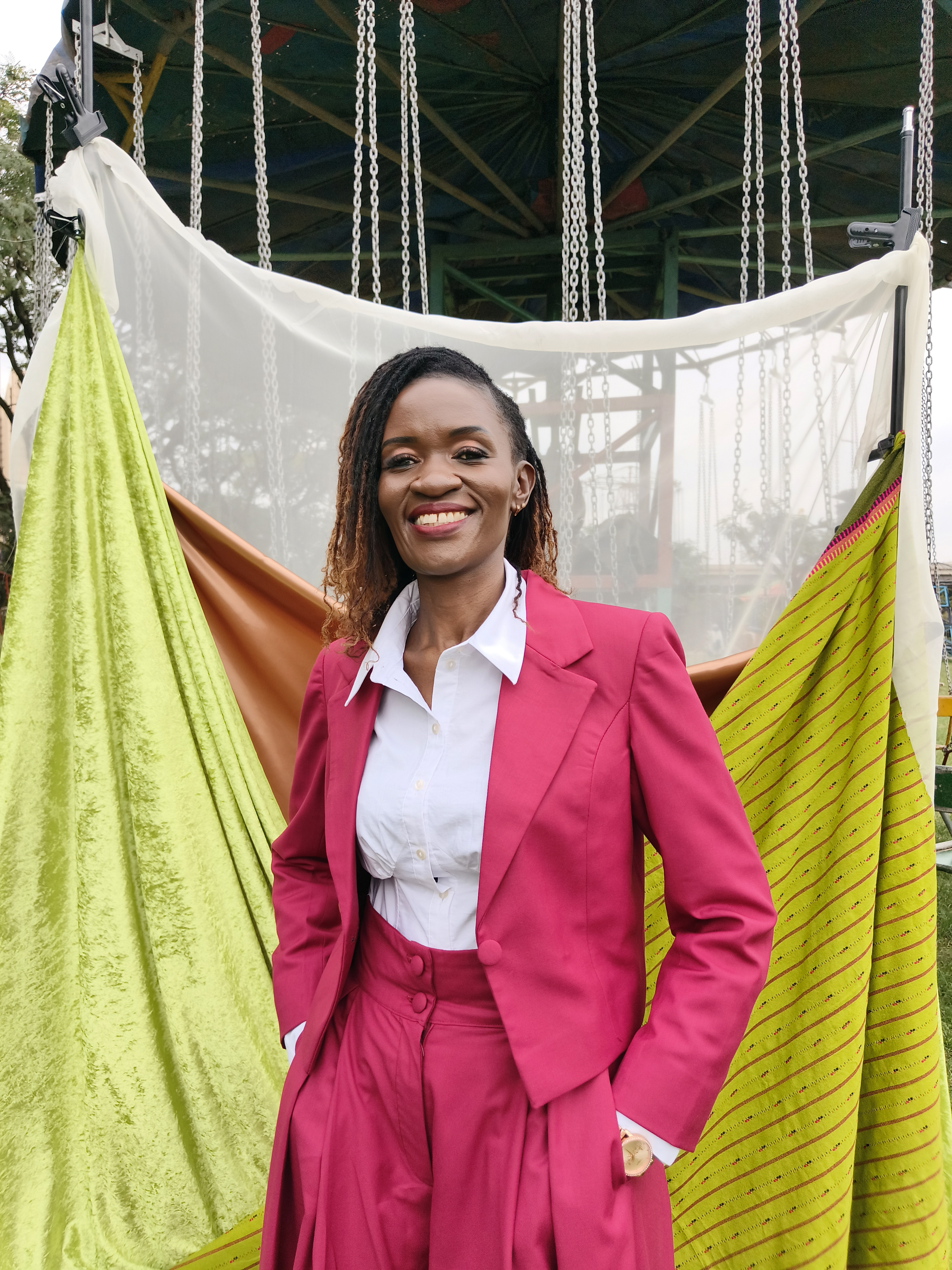A smiling woman with copper-tipped locs, wearing a raspberry trouser suit and white shirt, stands in front of lime-green and orange fabrics