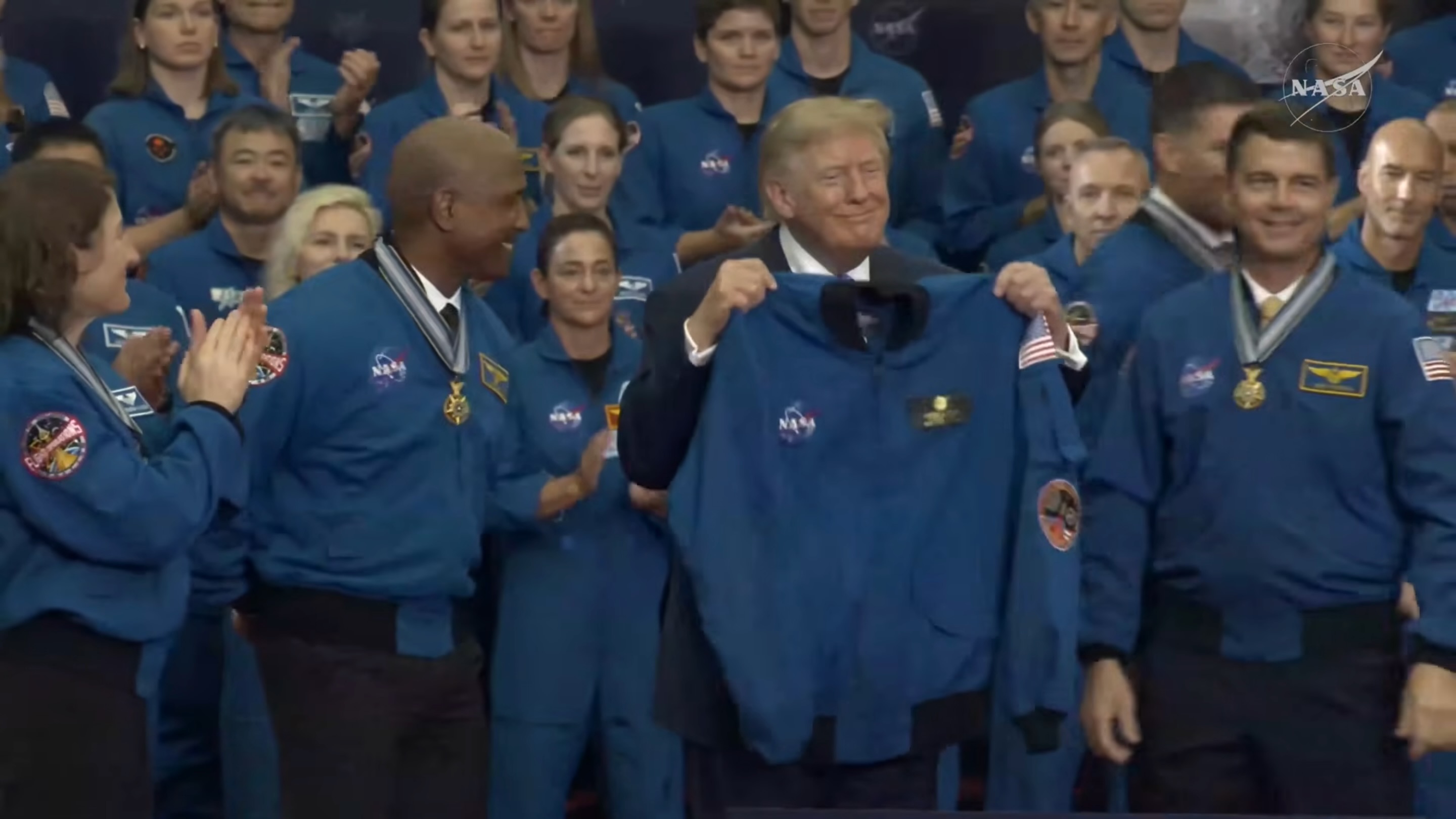 a smiling man in a dark suit holds up a blue flight jacket surrounded by a crowd of people wearing similar jackets