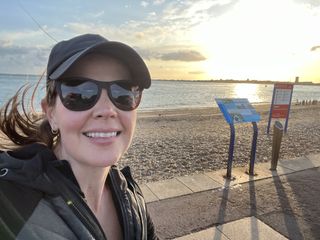 BBC weather presenter Alexis Green standing on a pebble beach wearing sunglasses, a baseball cap and a jacket