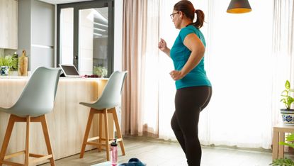 A woman marches on the spot at home. In front of her is a kitchen island with a laptop propped open.