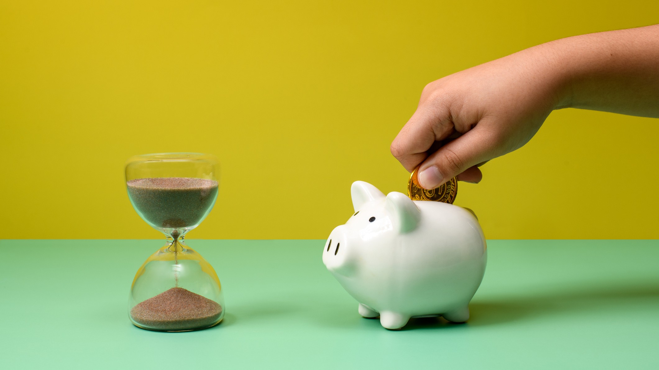 a hand deposits a coin into a piggy bank next to an hourglass