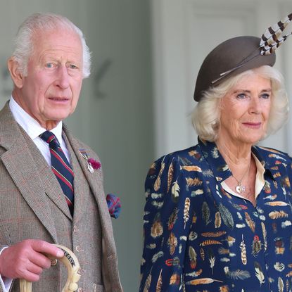 King Charles wears a a tweed suit and carries an umbrella while Queen Camilla wears a blue suit with a feather-print