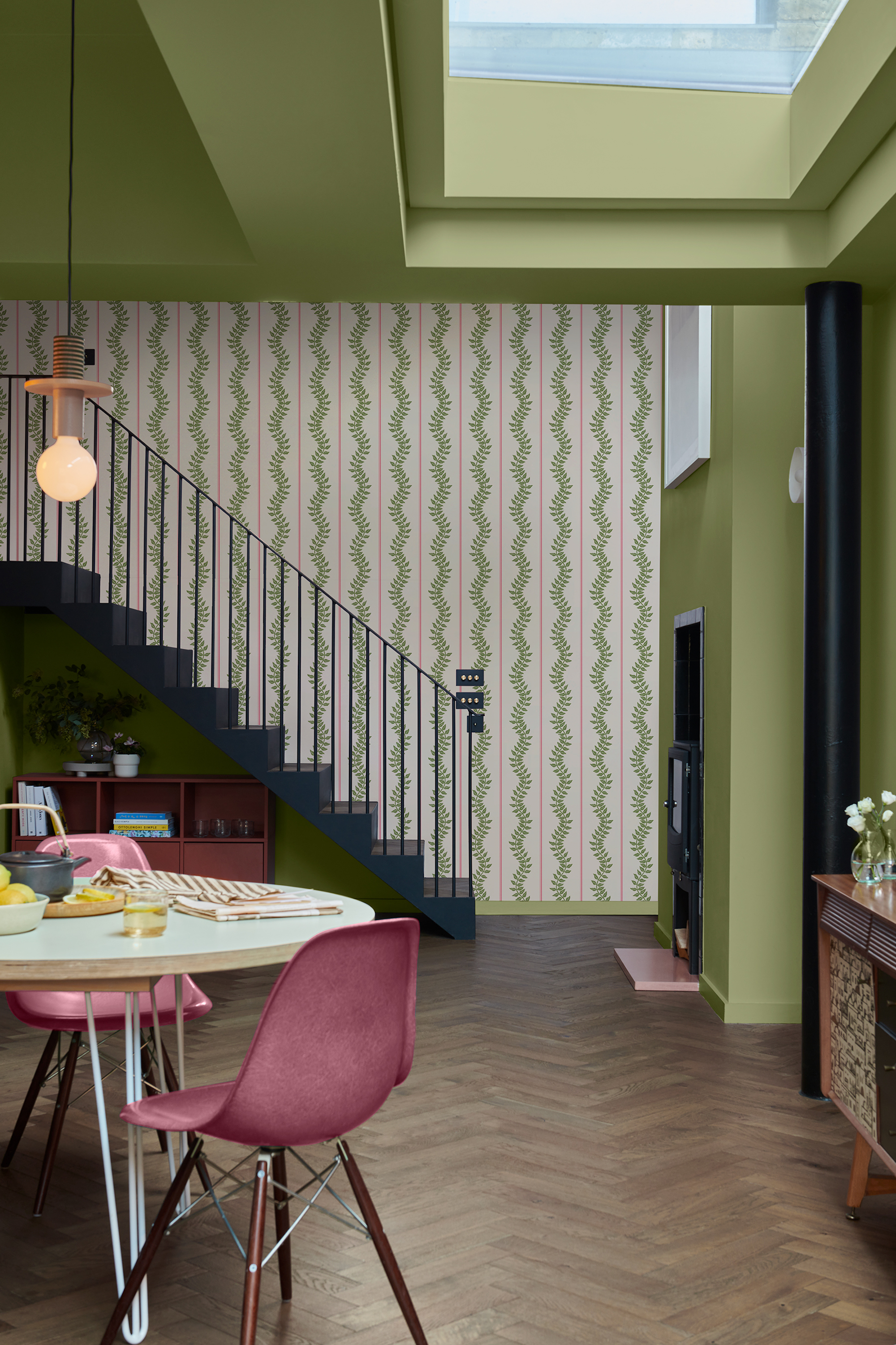 View into a sage green dining space, leading through to the stairs. The wall paper on the far wall is a striped pattern with a leafy design. The round dining table sits to the left of the space with a pendant light above.