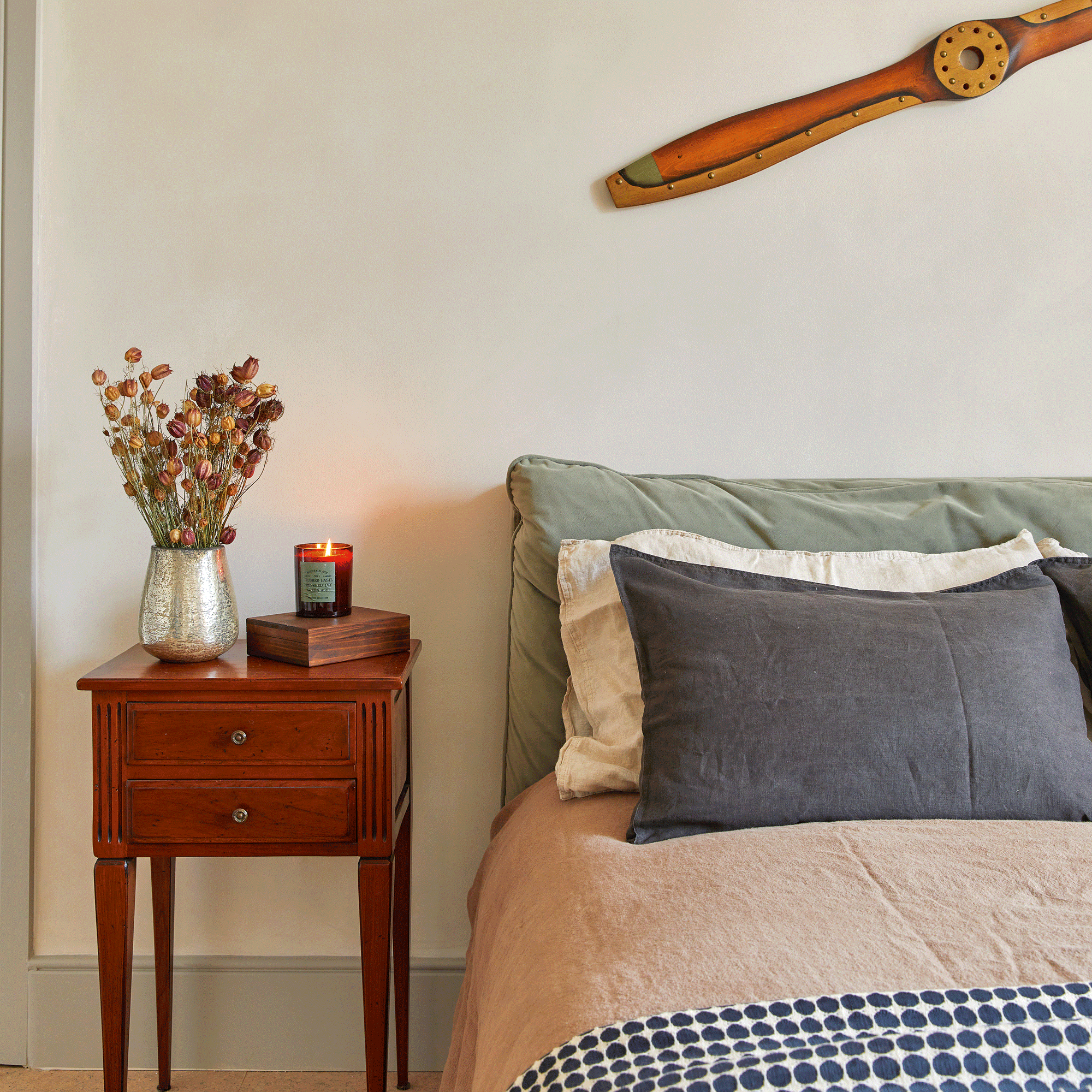 a bedroom with cream walls and a bed with layers of plain and patterned bedding and a vintage propellor on the wall
