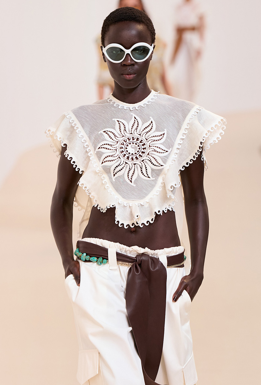 The coastal boho trend is shown in Zimmerman's spring summer 2025 runway show, with a model walking down the runway wearing oversized white sunglasses, a white eyelet flowy blouse with low-rise white trousers, a brown belt, and a turquoise chain belt