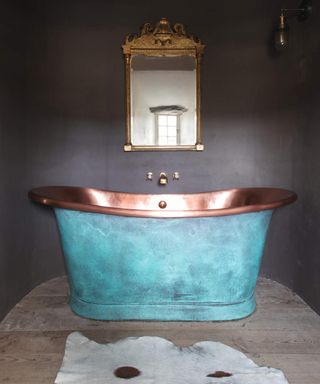 A bathroom with dark wall paint, animal print faux rug and copper bath painted with blue paint with ornate brass mirror design