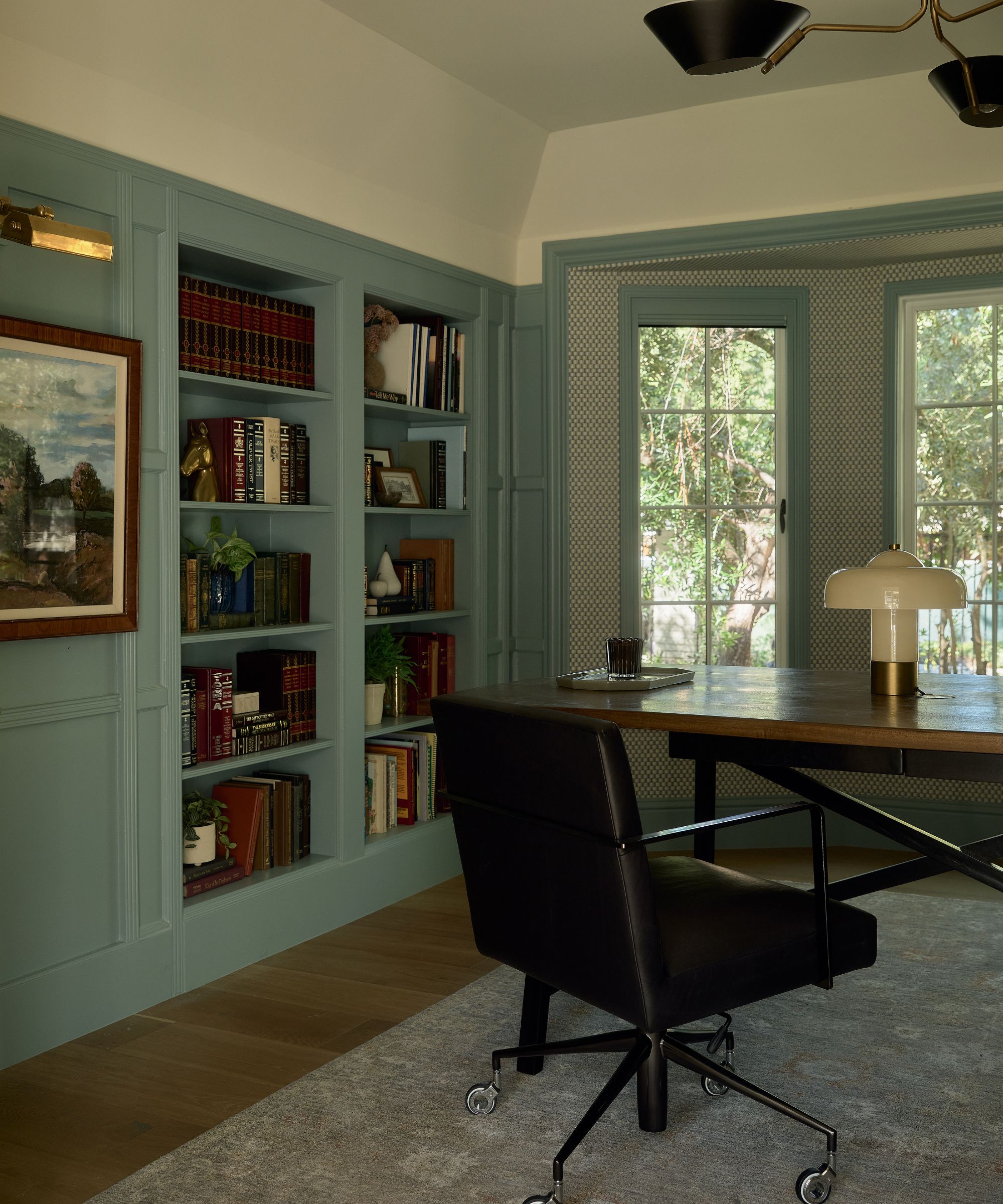 a blue painted paneled home office with a wallpapered bay window, built-in library and wooden office