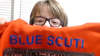 Blue Scuti holds up an orange hoodie with the words "Blue Scuti" and an image of the game crash proudly emblazoned on it.