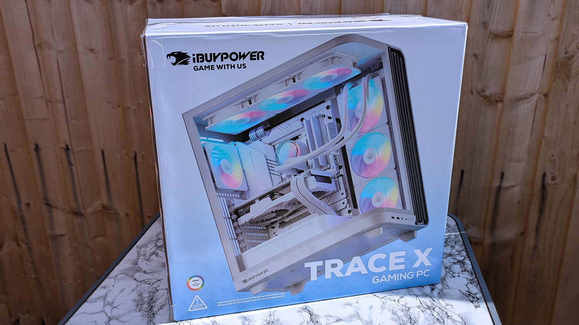 iBUYPOWER Trace X pre-built gaming PC in box, on a marble-effect table by a wooden fence
