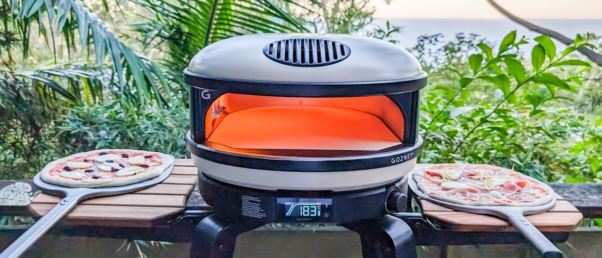 Gozney Arc XL pizza oven fired up on its stand with two uncooked pizzas on peels on fold-up shelves