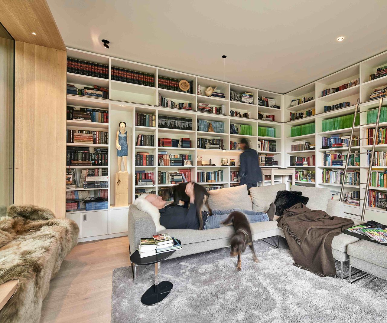 The library room of a contemporary self build built by Baufritz. The room features built-in bookcases on two wall and a large picture window. There is also a central sofa where the homeowner sits with his dog. The other homeowner looks at books in the background.